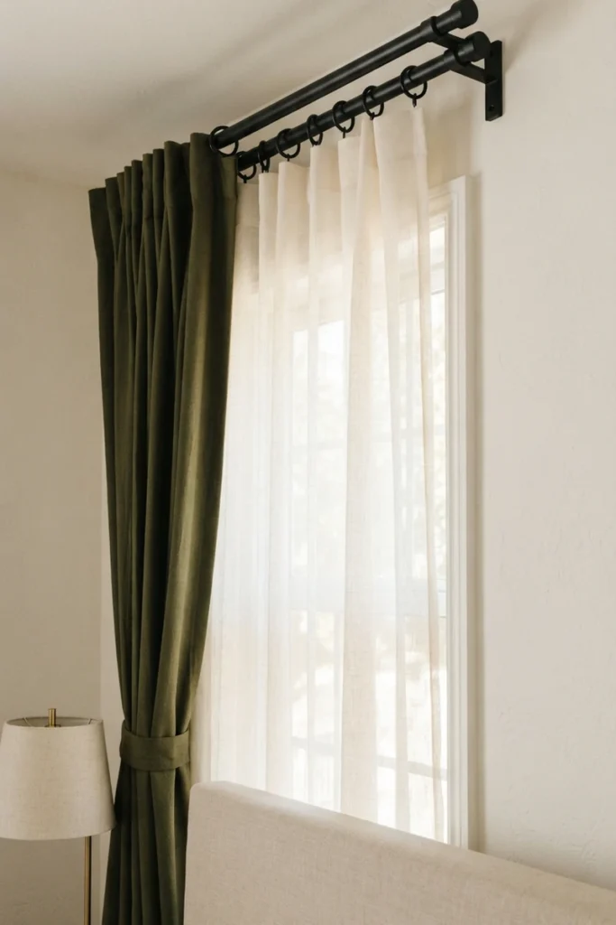 Bedroom blackout curtains in olive green and ivory sheers hanging on separate rings from a matte black double curtain rod